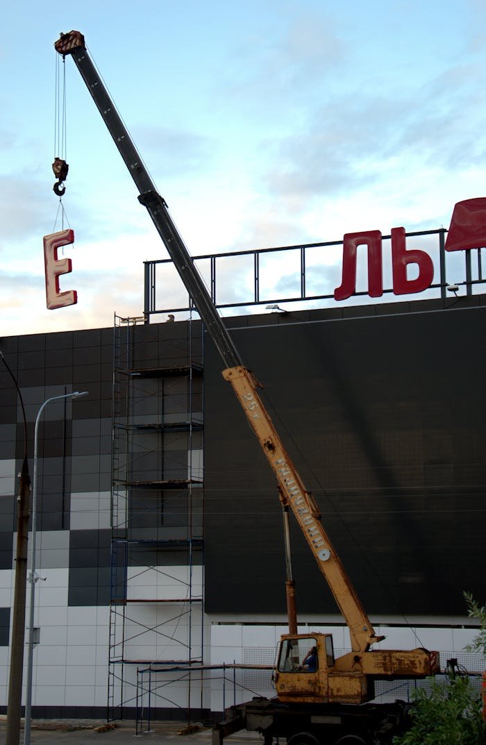 A construction crane lifting letter signage onto a building during daytime.
